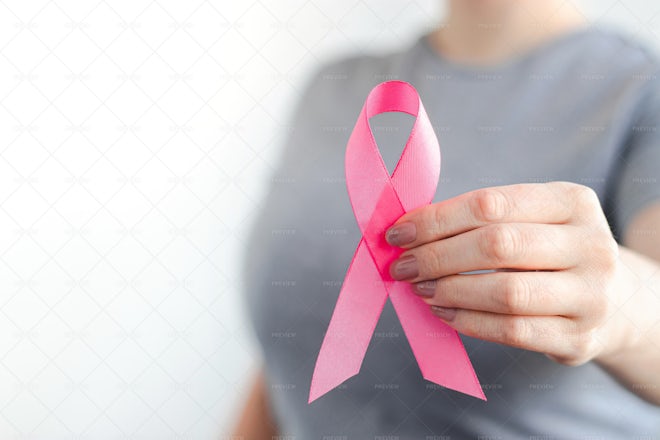 Woman hand holding a light pink ribbon on a pink background - cancer symbol  Stock Photo by wirestock