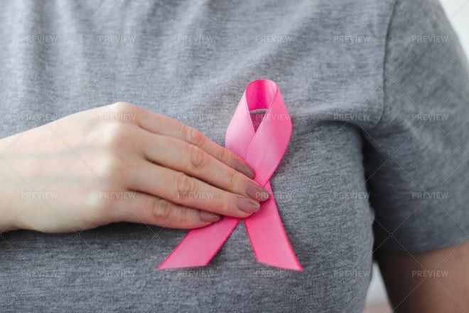 Woman hand holding a light pink ribbon on a pink background - cancer symbol  Stock Photo by wirestock